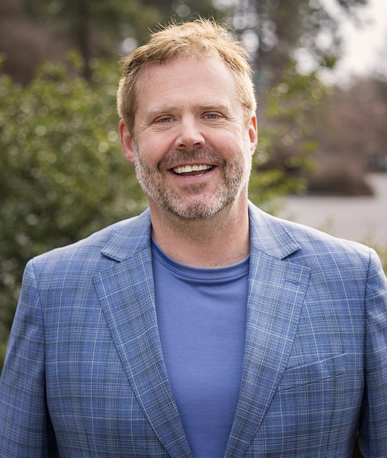 Person smiling outdoors in a blue plaid jacket, with trees in the background.