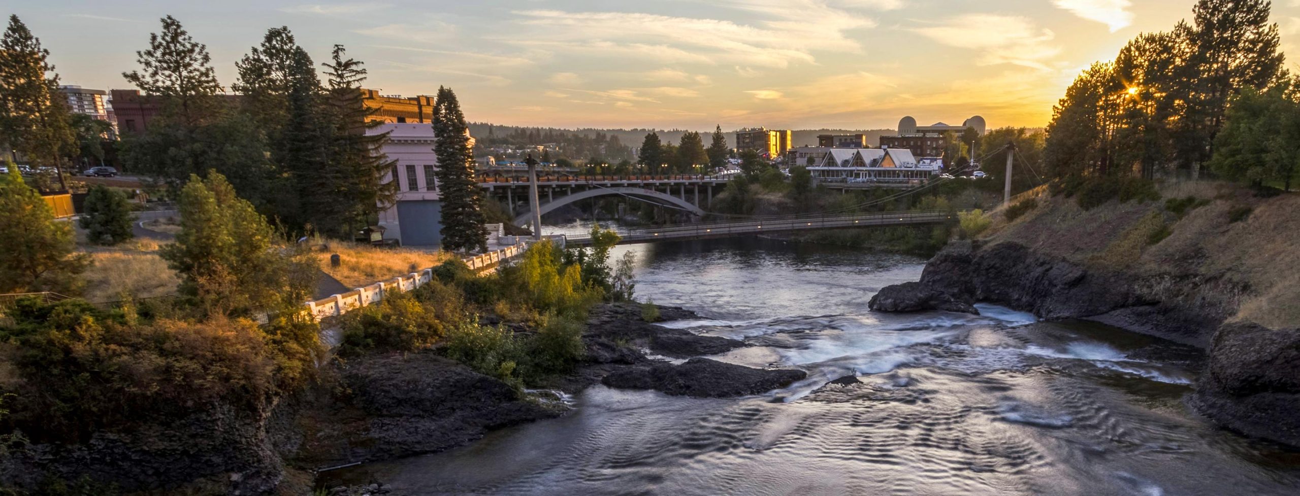 River flowing through a cityscape at sunset with a bridge in view.