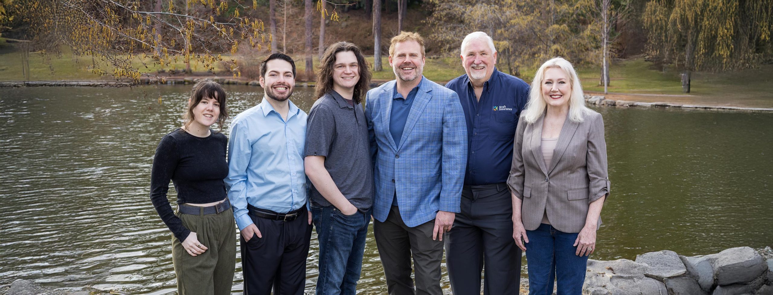 Team of six people standing together by a lake in a park setting.